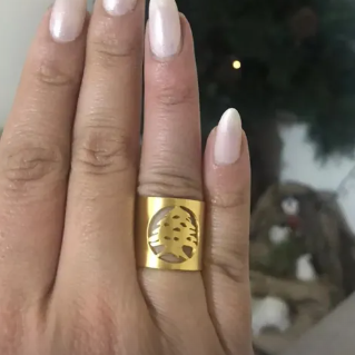 A close-up of a hand with long, glossy nude-pink manicured nails wearing a wide, chunky gold ring with a circular cutout design on the ring finger, set against a blurred Christmas tree background.