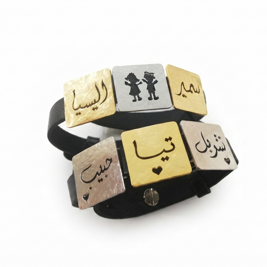 A black leather bracelet with five rectangular metal nameplates in gold and silver, featuring Arabic calligraphy and a small silver heart, shown on a white background.