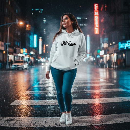 Woman in a white hoodie with Arabic text habibet 3amti standing on a city street at night.