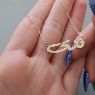 Gold necklace with Arabic script held in a hand against a neutral background