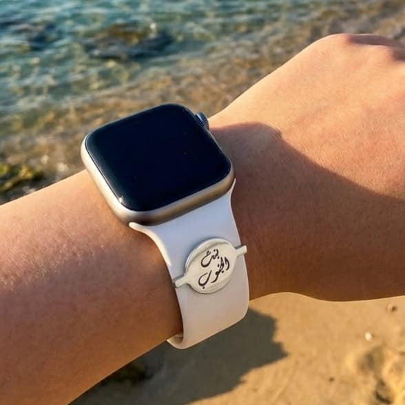 A close-up of a person's wrist wearing a smartwatch with a white band featuring a circular silver emblem with Arabic calligraphy, set against a blurred beach and ocean background.