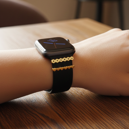 Smartwatch with a gold flower and heart designs on a black band on a wrist, set against a blurred indoor background.