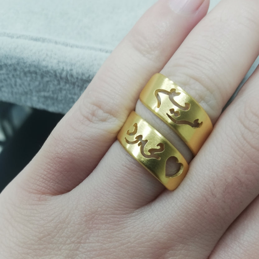 A close-up of a person's hand wearing two names on a rolled gold rings with Arabic calligraphy cutouts.