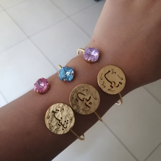 Four gold-tone bracelets with round engraved discs and colorful gemstone accents, shown worn on a person's wrist against a light tiled floor background.