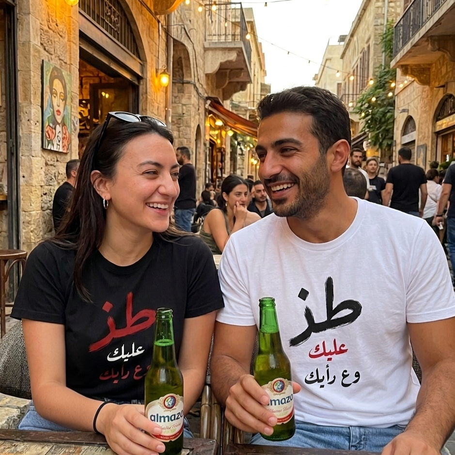 A smiling couple at an outdoor cafe table in a historic city street, holding green bottles. The man wears a white t-shirt with black Arabic calligraphy reading 'toz 3alek w 3a ra2yak, and the woman wears a black t-shirt with red Arabic calligraphy.