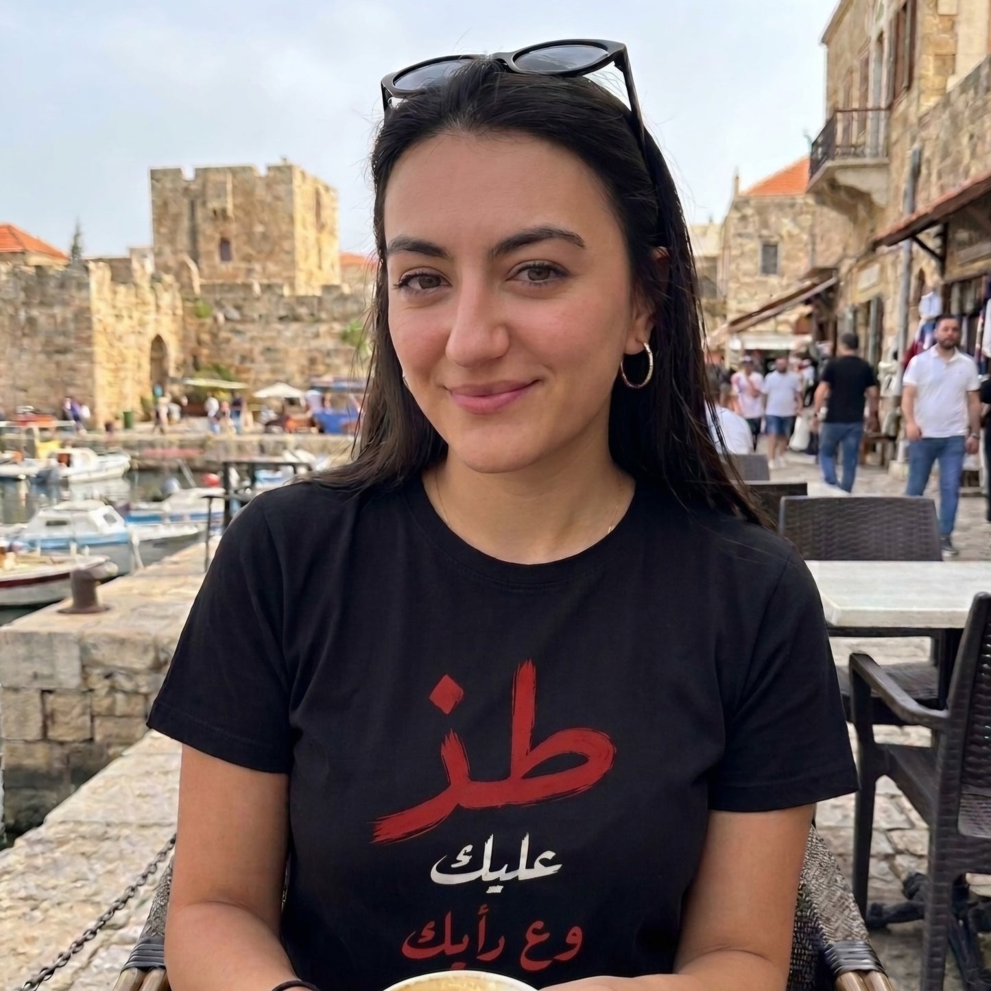 A woman wearing a black graphic t-shirt with red and white Arabic text, sitting outdoors in front of a stone harbor and historic building. Toz mind your business in arabic