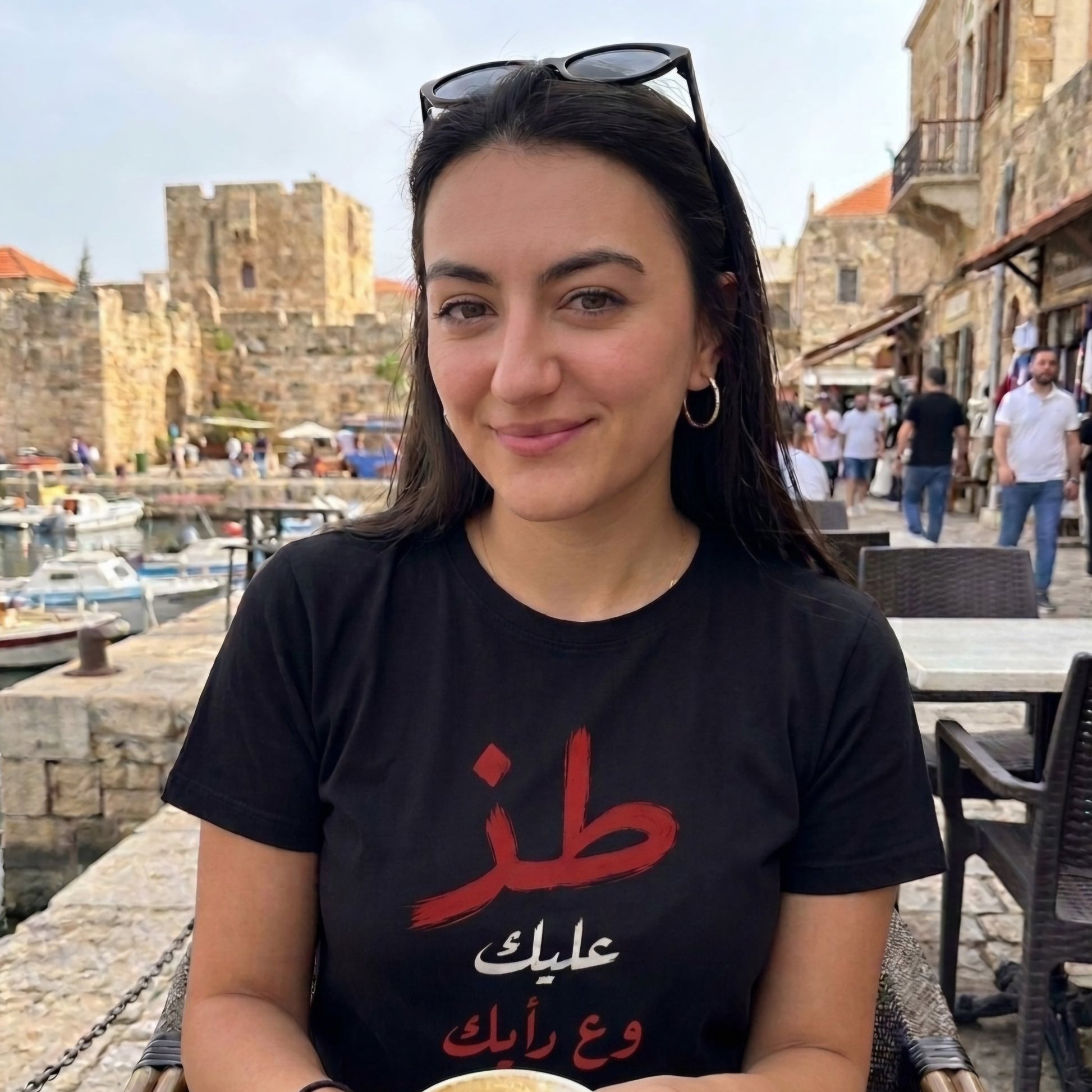 A woman wearing a black graphic t-shirt with red and white Arabic text, sitting outdoors in front of a stone harbor and historic building. Toz mind your business in arabic