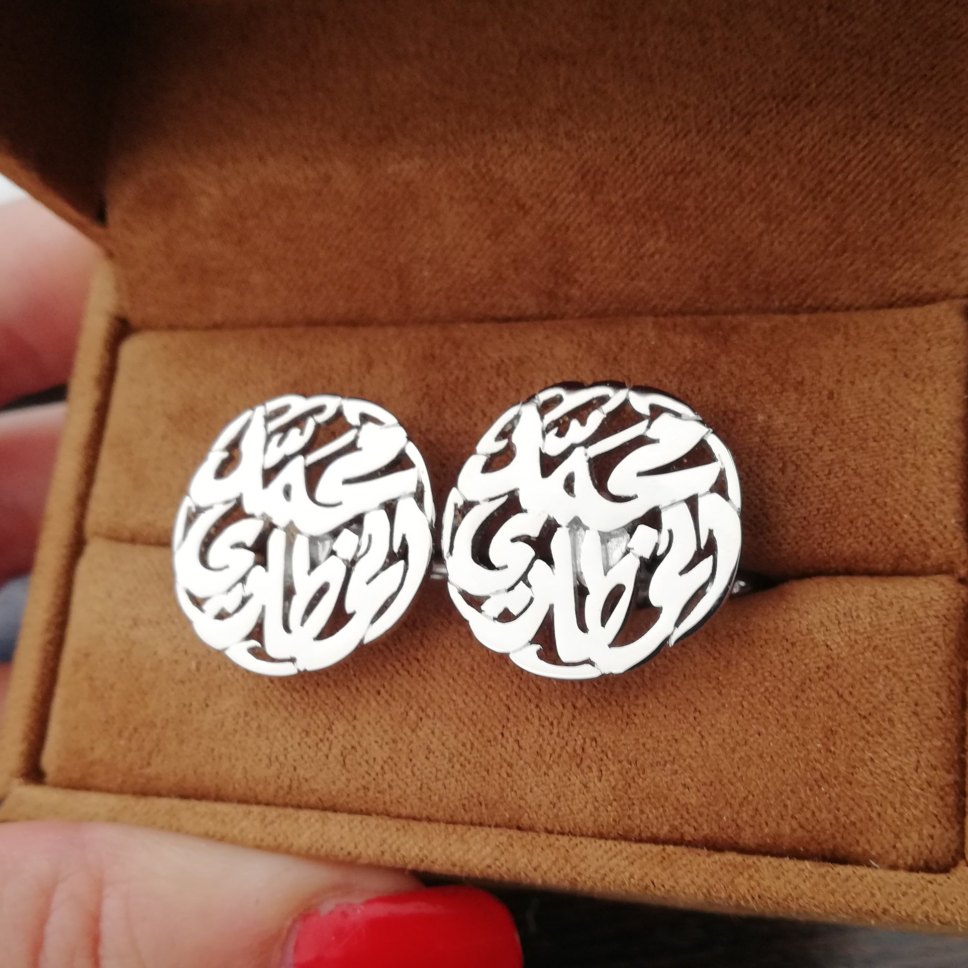 Silver cufflinks in a brown jewelry box held by a person with red nail polish.