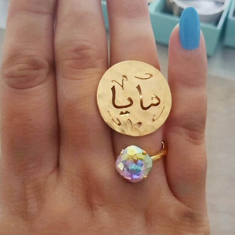 A close-up of a hand with blue nail polish wearing a gold ring with Arabic calligraphy with a changeant crystal color swarovski.