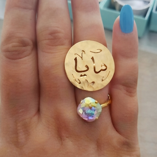 A close-up of a hand with blue nail polish wearing a gold ring with Arabic calligraphy with a changeant crystal color swarovski.