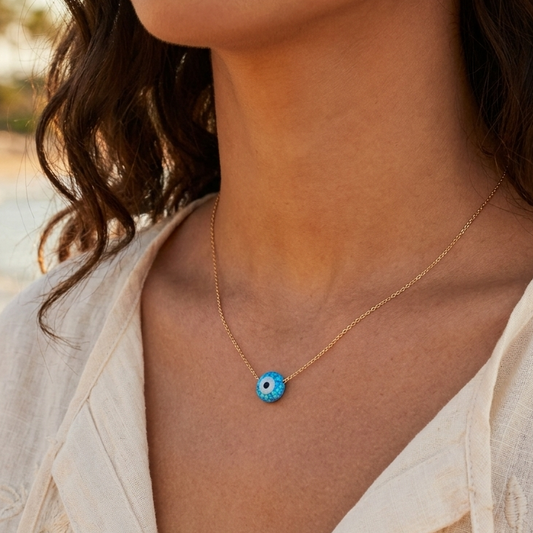 Woman wearing a gold necklace with a blue evil eye pendant on a blurred beach background