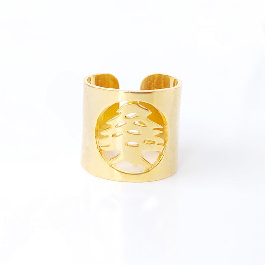 A gold-toned metal cuff ring featuring a circular cutout with a stylized tree design, set against a plain white background.