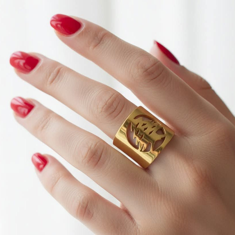 A close-up of a hand with red manicured nails wearing a wide, chunky gold ring with a lebanese cedar design