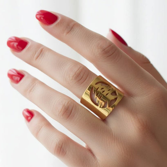 A close-up of a hand with red manicured nails wearing a wide, chunky gold ring with a lebanese cedar design