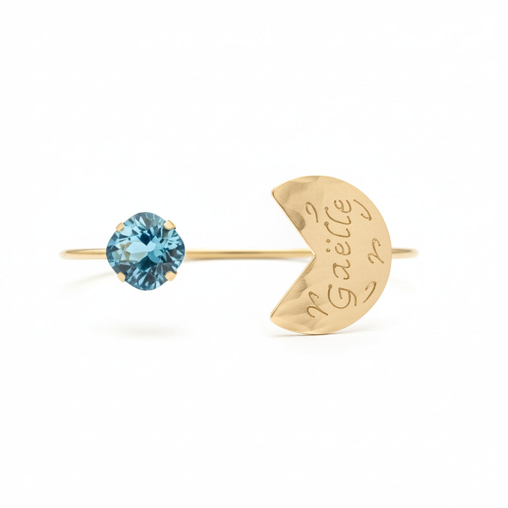 Gold bracelet with blue gemstone and heart-shaped tag on a red cushion against a gray background.