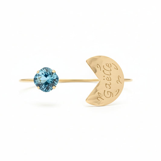 Gold bracelet with blue gemstone and heart-shaped tag on a red cushion against a gray background.