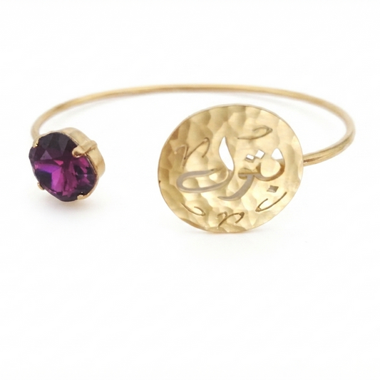 A gold-plated open cuff bracelet featuring a hammered circular disc engraved with Arabic calligraphy and a bezel-set round purple amethyst gemstone on one end, shown against a plain white background.