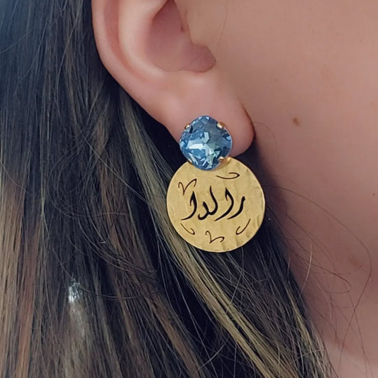 A close-up of a person's ear wearing a gold disc earring with Arabic calligraphy and a blue cushion cut swarovski.