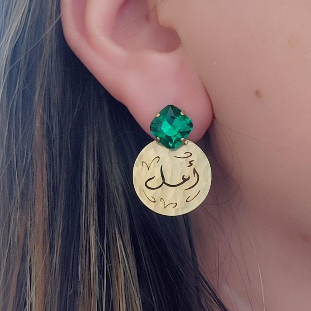 A close-up of a person's ear wearing a gold-tone circular drop earring with black Arabic calligraphy and a faceted light blue gemstone at the top.