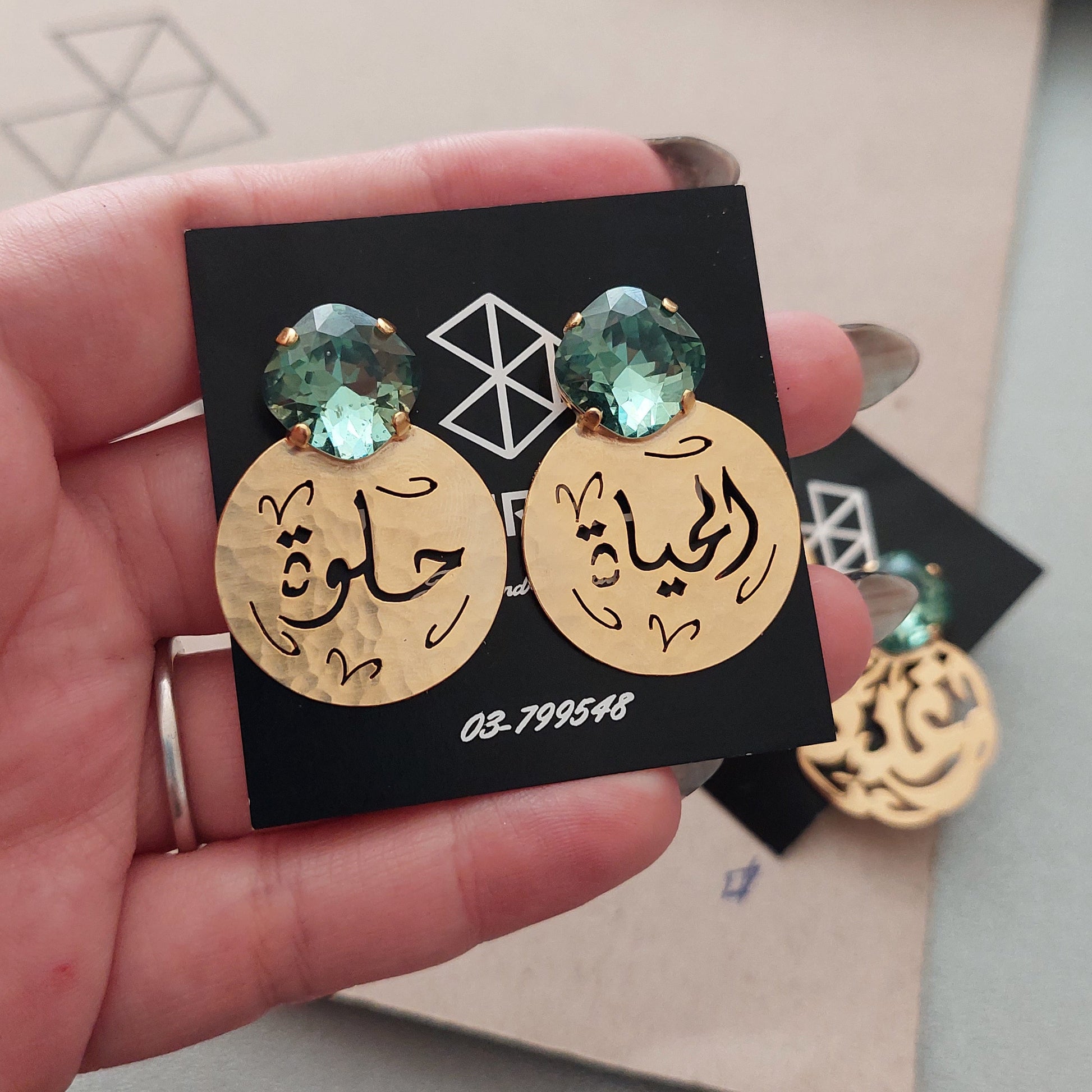 A hand holding a black display card featuring two gold-toned earrings with engraved face designs and green erinite swarovski, with more earrings visible in the background.