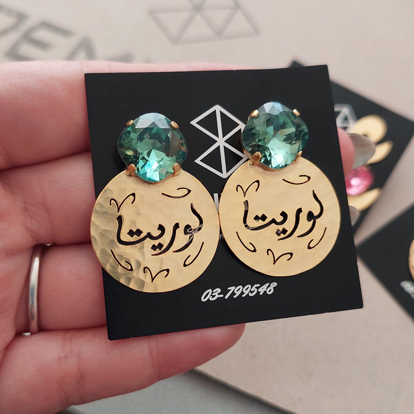 A hand holding a black display demirelli card featuring a pair of gold-tone stud earrings with Arabic calligraphy and erinite swarovski, with a beige geometric box in the background.
