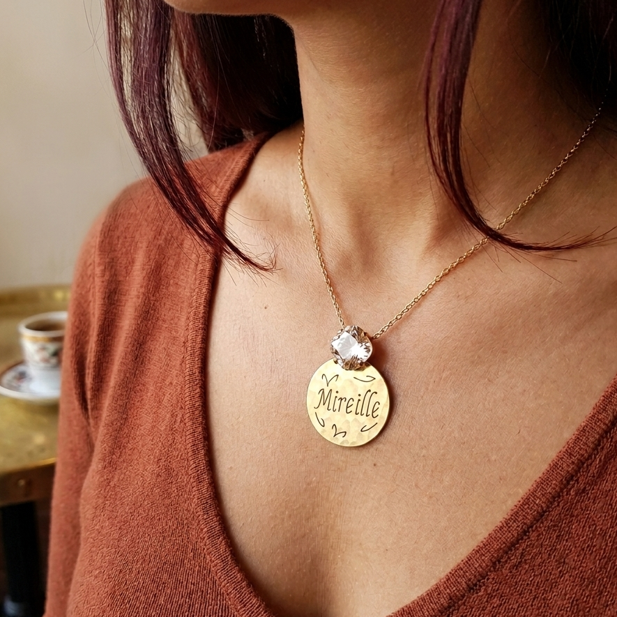 A close-up lifestyle shot of a woman wearing a gold pendant necklace with a clear faceted gemstone and a round gold charm engraved with the name 'Mireille'. She is wearing a rust-brown V-neck top, with a warm indoor background featuring a table and cup.