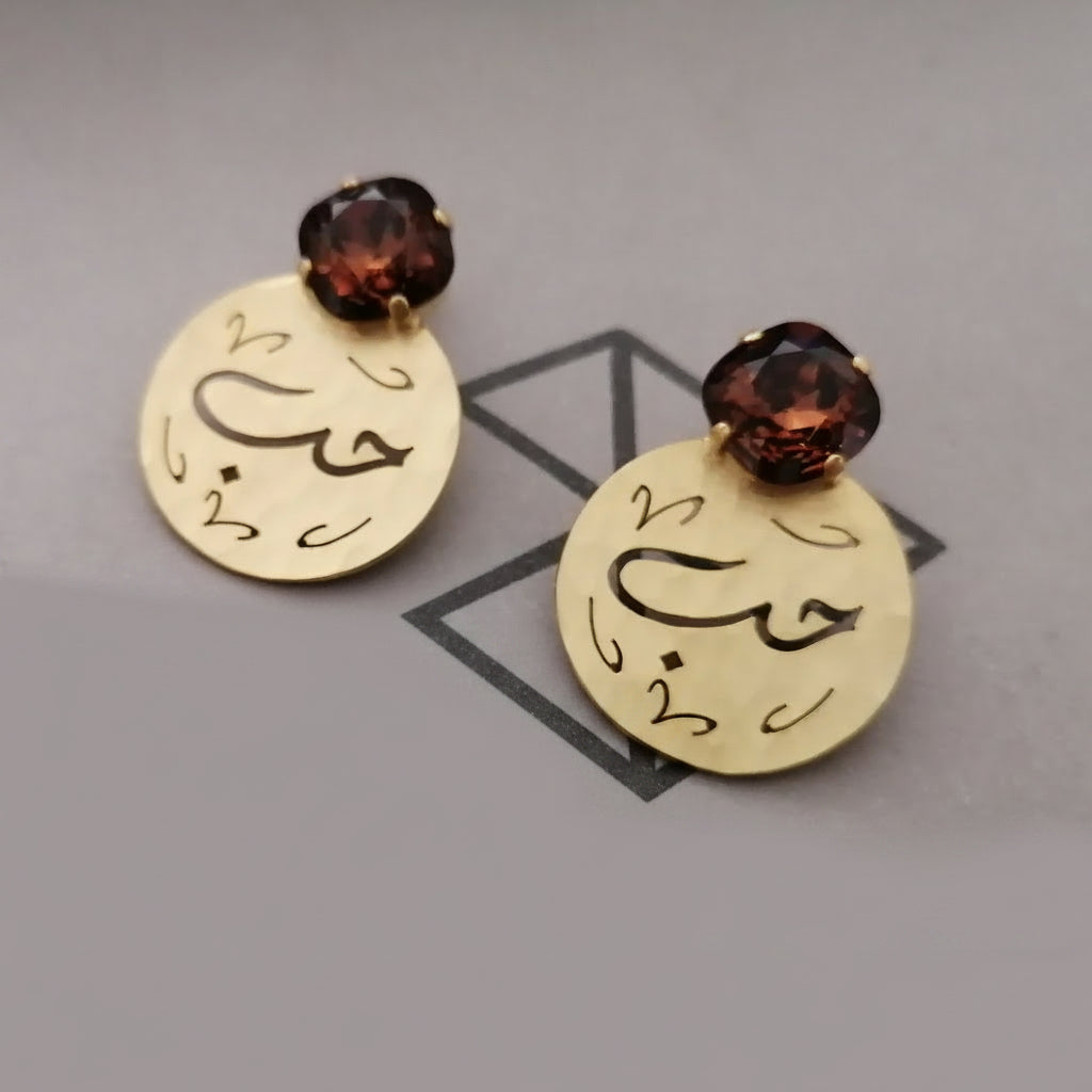 A pair of gold-toned drop earrings with round engraved discs featuring Arabic calligraphy, topped with faceted dark smoke topaz swarovski , displayed on a gray surface.