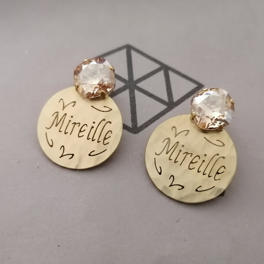 A pair of gold-toned disc earrings engraved with the name 'Mireille' and topped with clear faceted rhinestones, displayed on a gray surface with a black geometric logo.