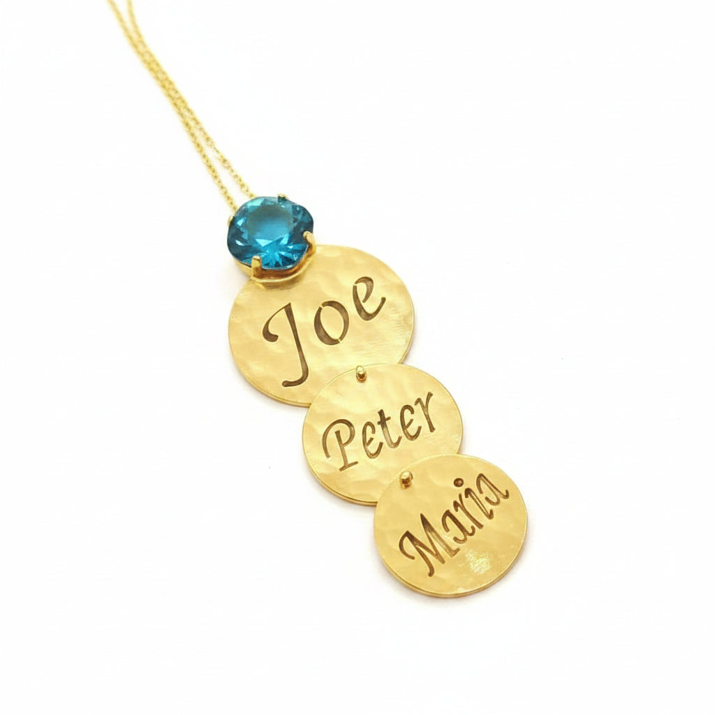 A gold-toned personalized necklace with three stacked engraved name discs reading 'Joe', 'Peter', and 'Maria', hanging from a gold chain with a small round blue swarovski.