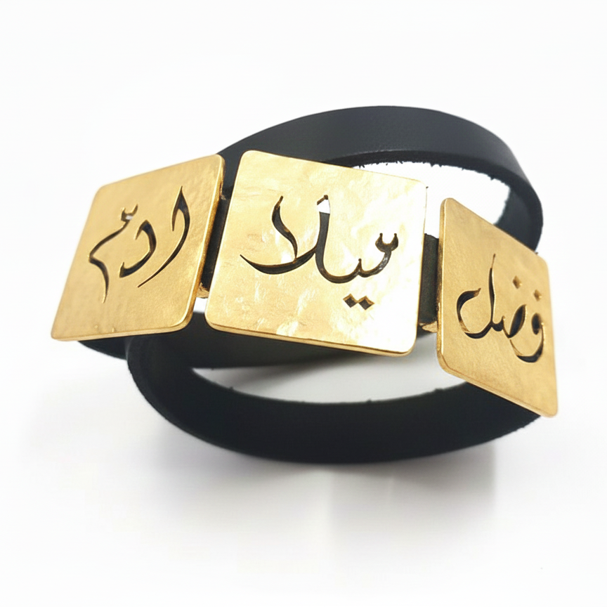 A black leather bracelet with three gold square plates engraved with Arabic calligraphy, displayed on a clean white background.