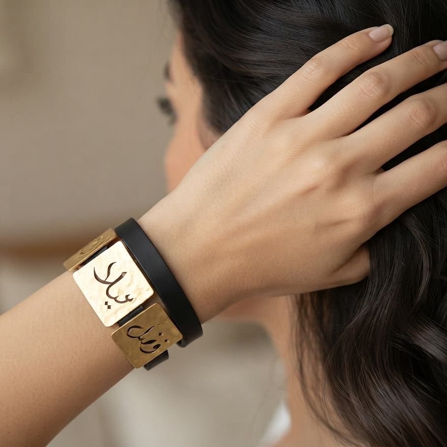 A close-up lifestyle shot of a woman wearing a black and gold Arabic nameplate cuff bracelet, with her hand resting on her dark hair.