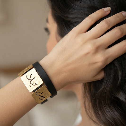 A close-up lifestyle shot of a woman wearing a black and gold Arabic nameplate cuff bracelet, with her hand resting on her dark hair.