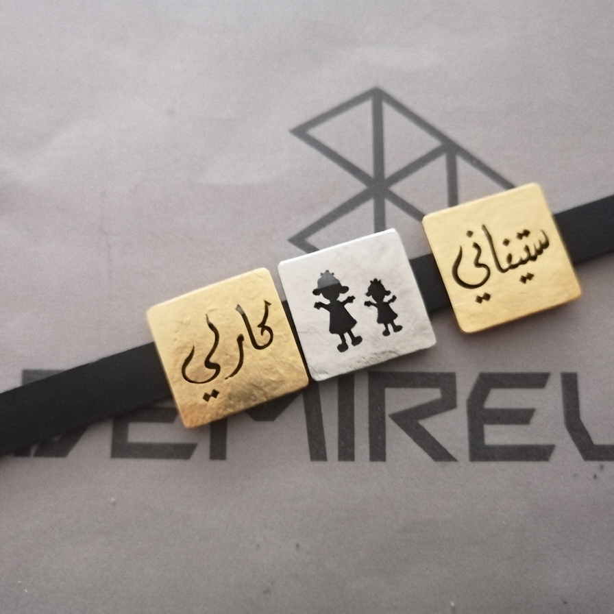 A black bracelet or band laid diagonally across a gray branded surface, featuring three square decorative charms: two gold-toned with black script and one silver-toned with two small mother and daughter figures.