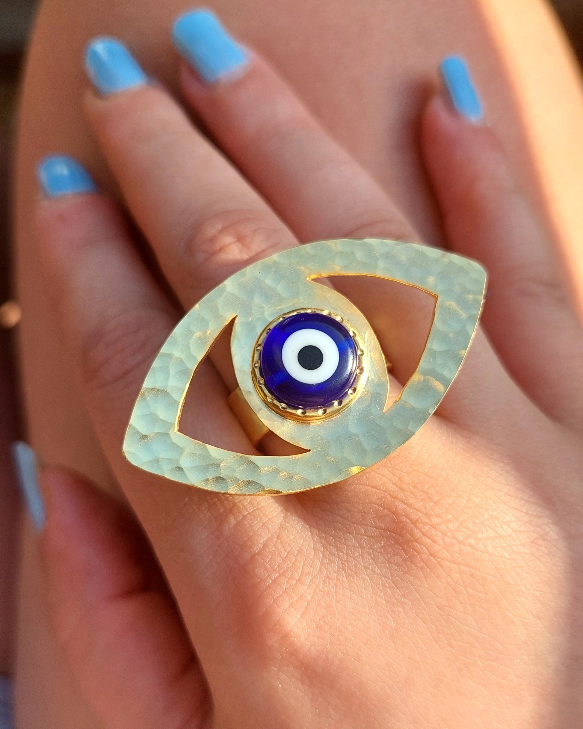 Gold eye-shaped ring with a blue center on a hand with light blue nail polish.
