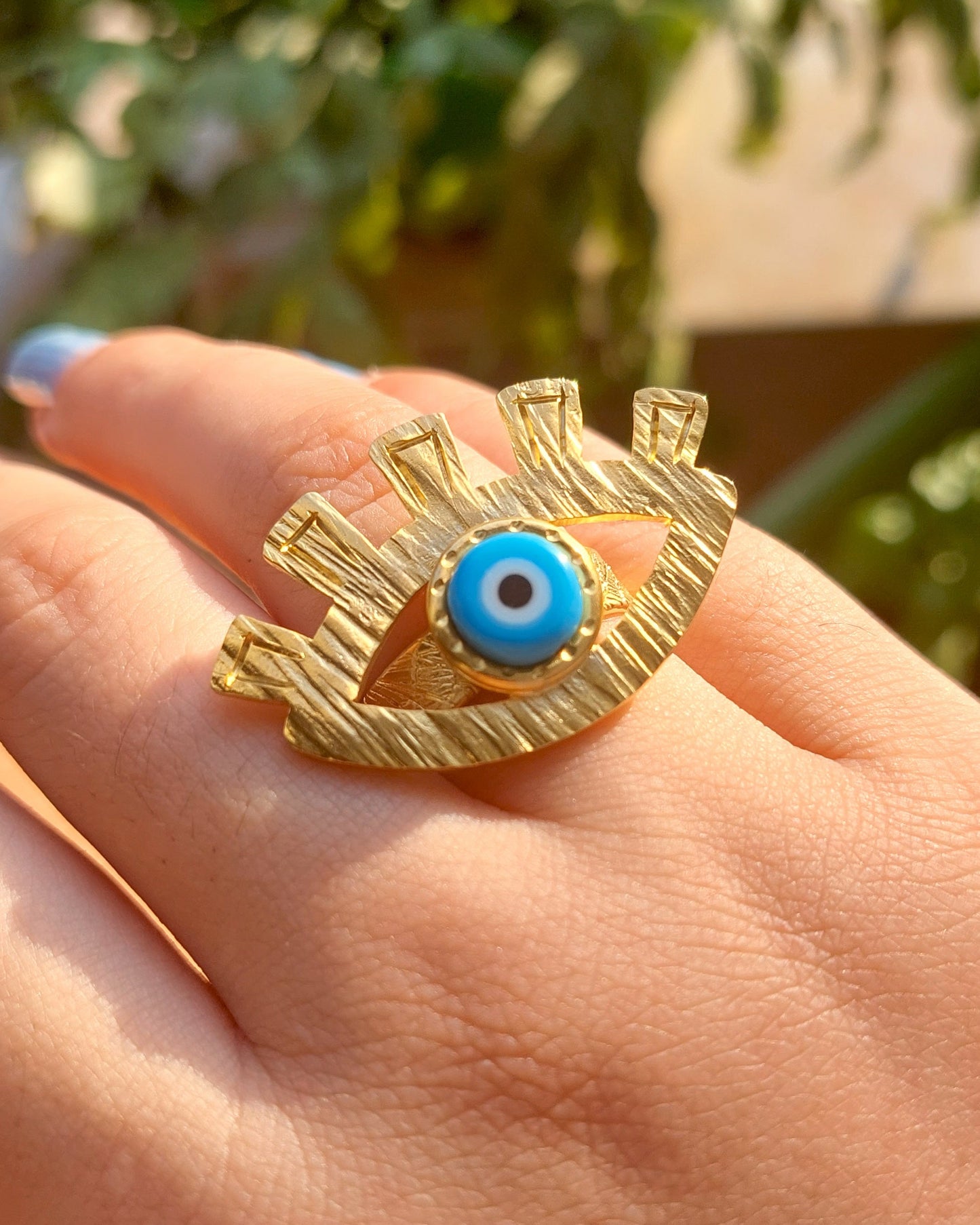 Gold ring with a blue eye design on a hand against a blurred green background