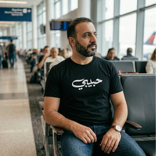 Man sitting in an airport terminal wearing a black t-shirt with Arabic text.