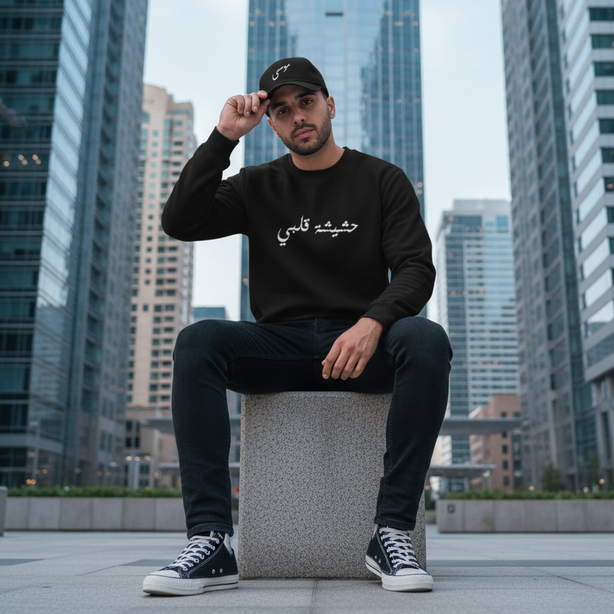 Man sitting on a block in an urban setting wearing a black sweatshirt with Arabic text written the weed of my heart.