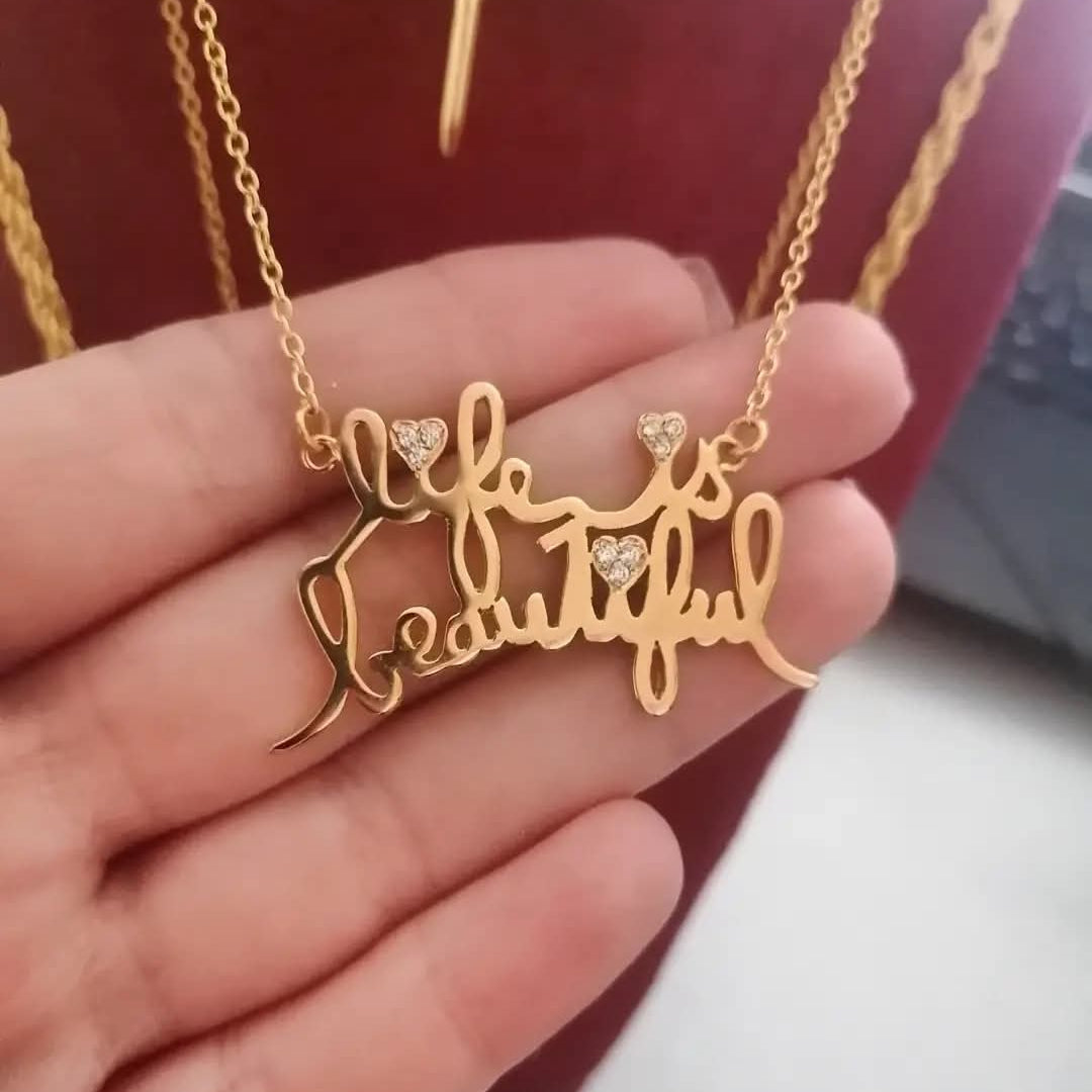 Gold necklace with 'life is beautiful' pendant held in a hand against a blurred background