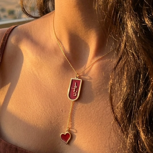 Close-up of a necklace with a red enamel name and red enamel heart pendant worn by a person against a blurred background.
