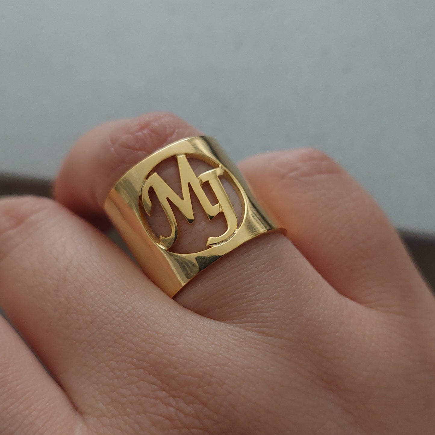 A close-up of a hand wearing a wide gold ring with a stylized cutout letter design.