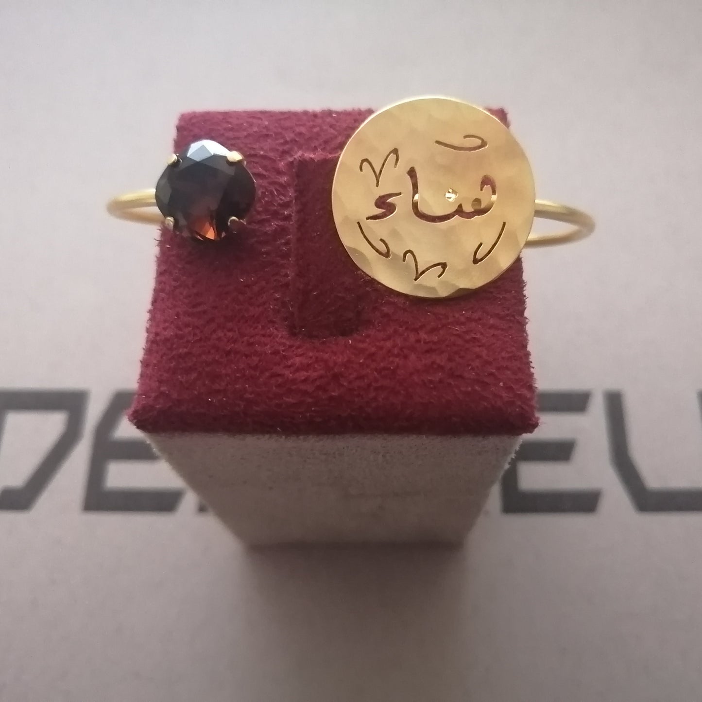 A gold bracelet hammered gold disc on the band with Arabic calligraphy engraved on it, alongside a small dark gemstone cluster.