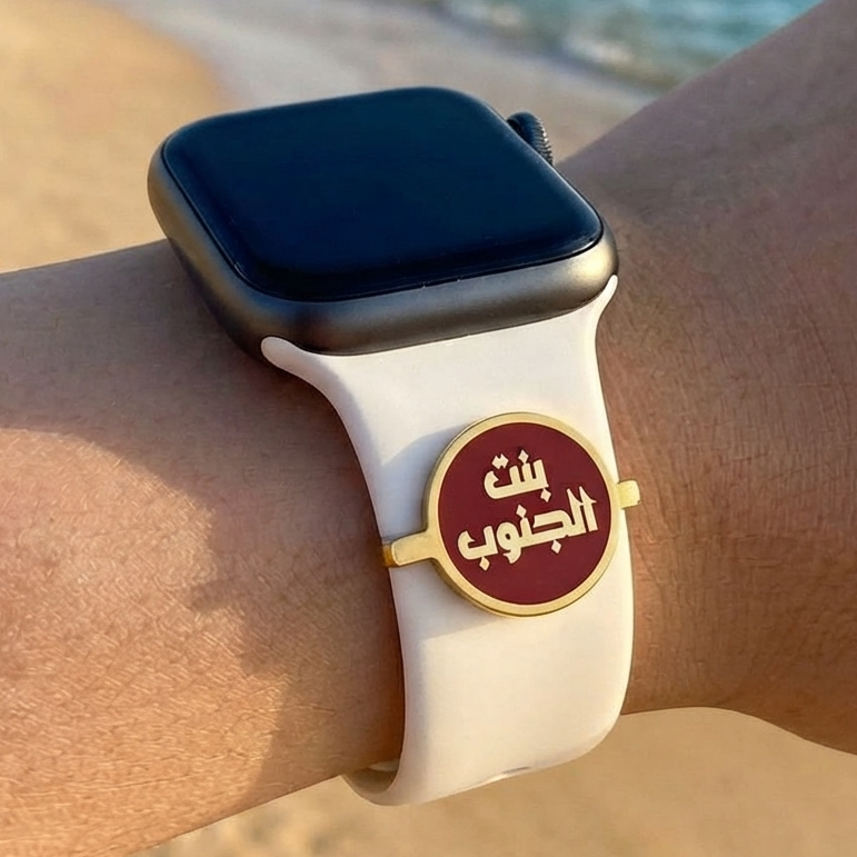 A close-up of a person's wrist wearing a smartwatch with a white sport band and a round gold-tone charm featuring Arabic calligraphy on a beach with the ocean in the background.