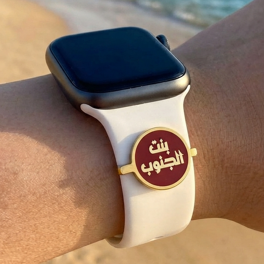 A close-up of a person's wrist wearing a smartwatch with a white sport band and a round gold-tone charm featuring Arabic calligraphy on a beach with the ocean in the background.