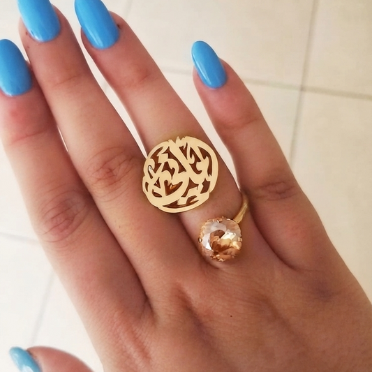 A gold ring with an arabic circular calligraphy name design and a small golden shadow cushion cut swarovsku, worn on a hand with bright blue nail polish against a light tiled background.