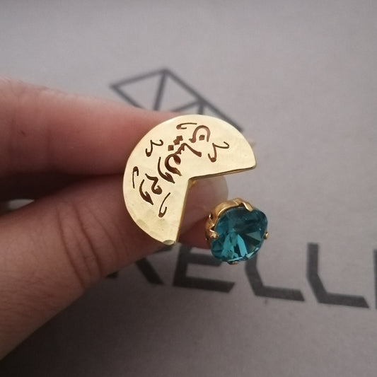 A gold crescent moon ring with engraved script and a small round teal blue gemstone, held between fingers against a gray background 