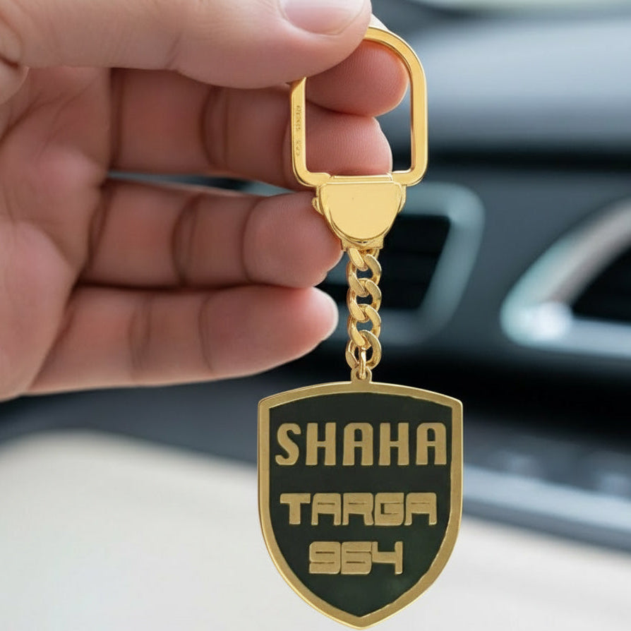 Gold keychain with 'SHAHA TARGA 954' text held by a hand in front of a car interior.
