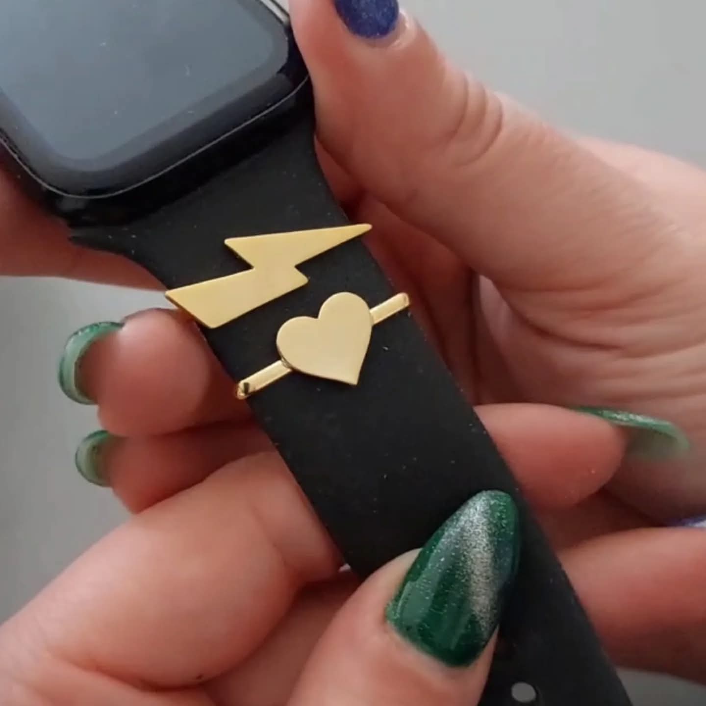 Person holding a smartwatch with a black strap featuring gold heart and lightning bolt designs.