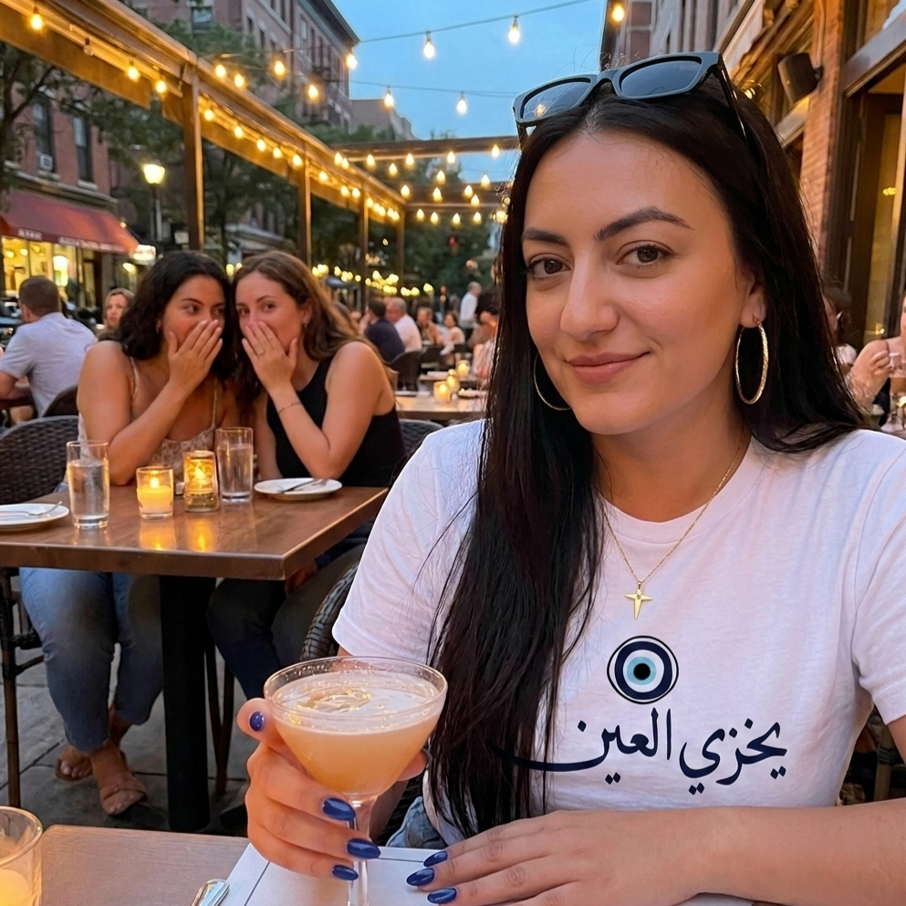 A woman at an outdoor restaurant table wearing a white t-shirt with Arabic yekhzi el 3ein text and an evil eye graphic. She is holding a cocktail and wearing gold hoop earrings and a necklace with a cross pendant.