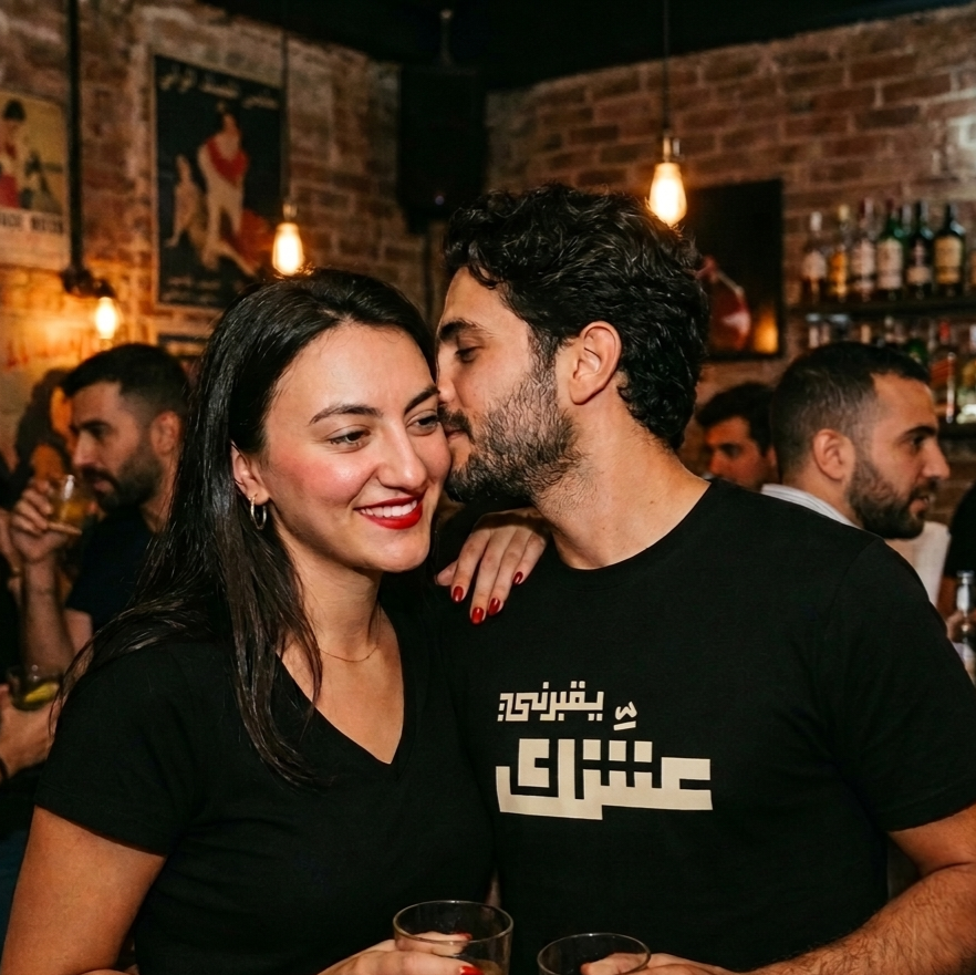 A man and a woman wearing matching black graphic t-shirts with white Arabic yo2borni 3eshik text, standing together in a lively, dimly lit bar with brick walls and other patrons in the background.