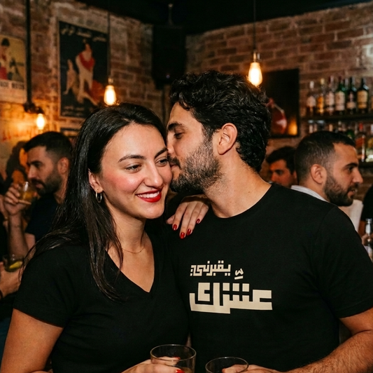A man and a woman wearing matching black graphic t-shirts with white Arabic yo2borni 3eshik text, standing together in a lively, dimly lit bar with brick walls and other patrons in the background.